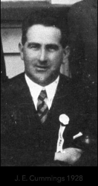 Historical black-and-white portrait of a man wearing a suit, striped tie, and a white rosette on his lapel.