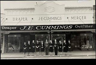 Black-and-white historical photograph of staff standing outside the "J. E. Cummings" drapery, mercer, and outfitter store on Arawa Street.