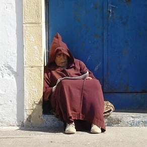 Moroccan man in a dark red hooded robe seated beside a blue doorway