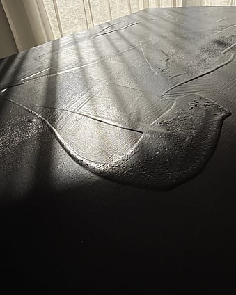 second detail image showing a warm black acrylic and silica sand surface in hazy sunlight, with glossy pooled forms, raised ridges and pitted texture with variations in sheen revealed by soft diffused light