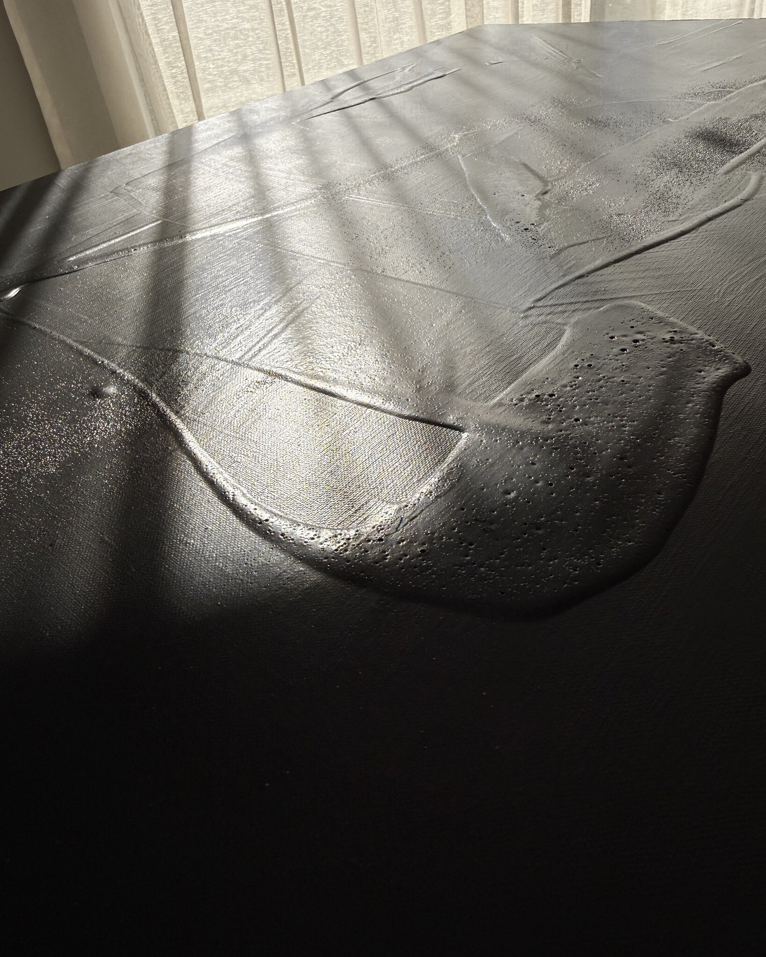 second detail image showing a warm black acrylic and silica sand surface in hazy sunlight, with glossy pooled forms, raised ridges and pitted texture with variations in sheen revealed by soft diffused light