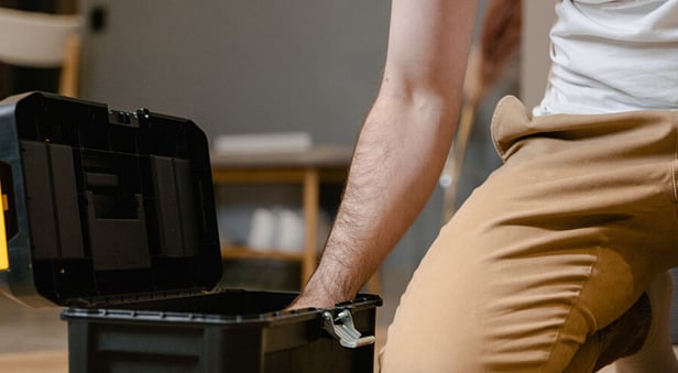 A person kneeling on a wooden floor reaching into an open black and yellow toolbox, with a tray of hand tools in the foreground.