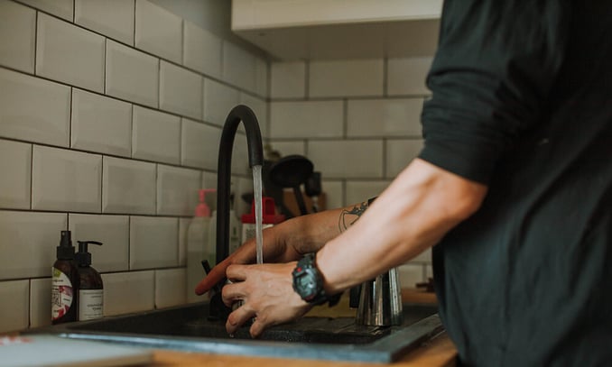 A person washing their hands under running water from a modern black faucet in a dark kitchen sink, set against a white subway tile backsplash.