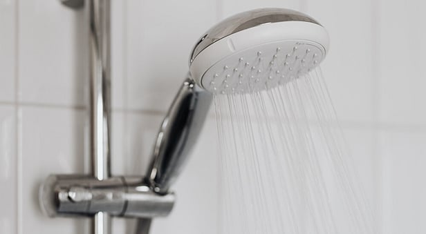 A close-up of a modern chrome showerhead spraying running water, mounted on a metal slide bar against a white square-tiled bathroom wall.