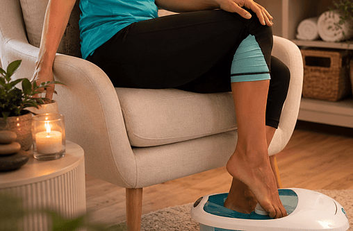 a lady having a foot spa sitting in a cream chair with a candle and plant on a table next to her
