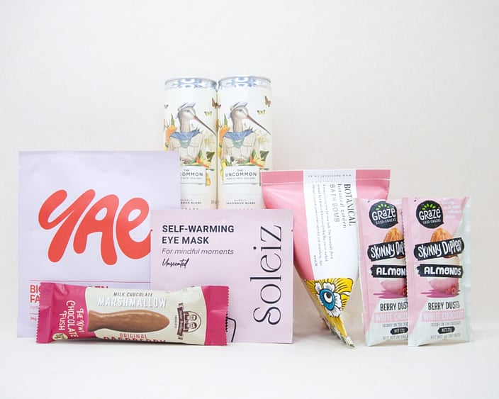 A Little Pick Me Up Gift Box featuring Uncommon sparkling wine, Soleiz eye mask, YAE face mask, Botanicals bath bomb, chocolate fish, and gourmet nuts, beautifully packaged as a self-care gift box under $100.