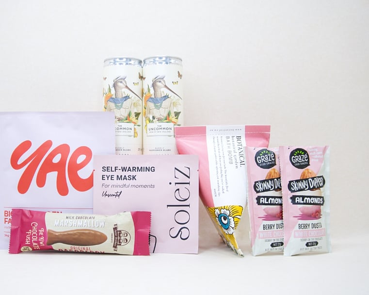 A Little Pick Me Up Gift Box featuring Uncommon sparkling wine, Soleiz eye mask, YAE face mask, Botanicals bath bomb, chocolate fish, and gourmet nuts, beautifully packaged as a self-care gift box under $100.