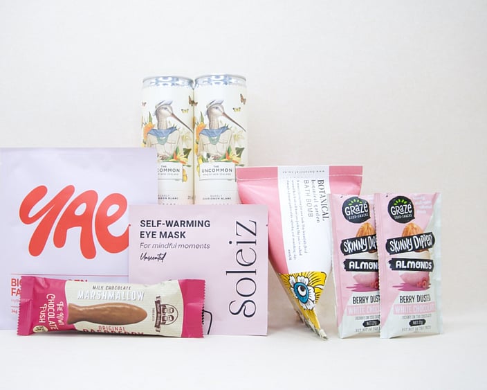 A Little Pick Me Up Gift Box featuring Uncommon sparkling wine, Soleiz eye mask, YAE face mask, Botanicals bath bomb, chocolate fish, and gourmet nuts, beautifully packaged as a self-care gift box under $100.
