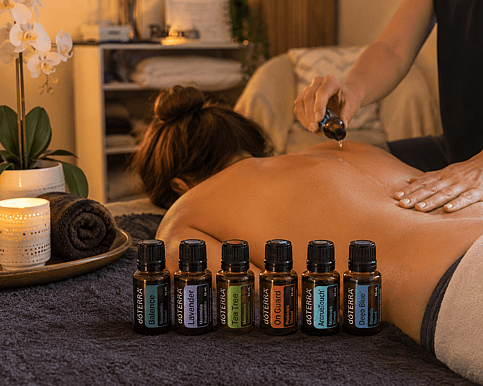 Picture of a lady lying face down having essnetial oils place on her back by the therapist. 8 essential oils are in front of her that will be used for the massage
