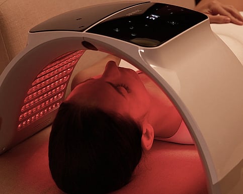 A picture of a lady lying on her back and has the led face light therapy pod over her head producing red light treatment