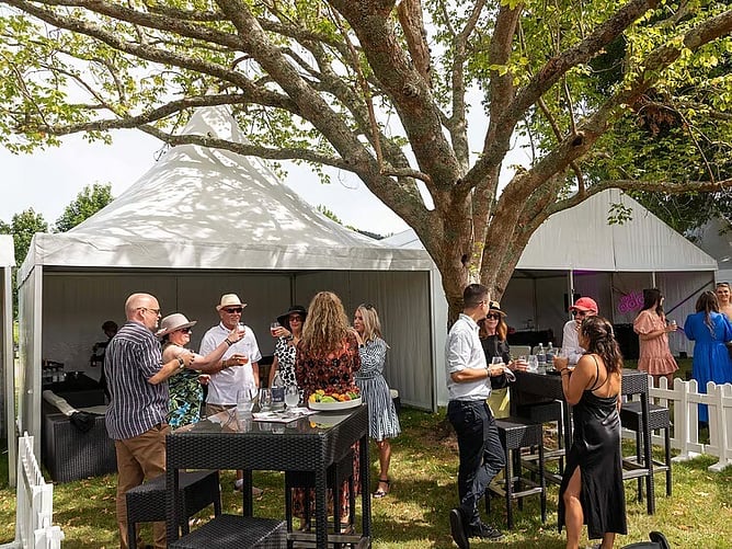 Guests gathered around outdoor bar tables beside a white marquee in a garden
