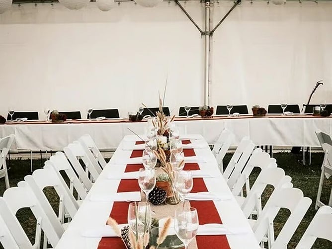 Long dining tables with white chairs, tablecloths and red runners inside a marquee