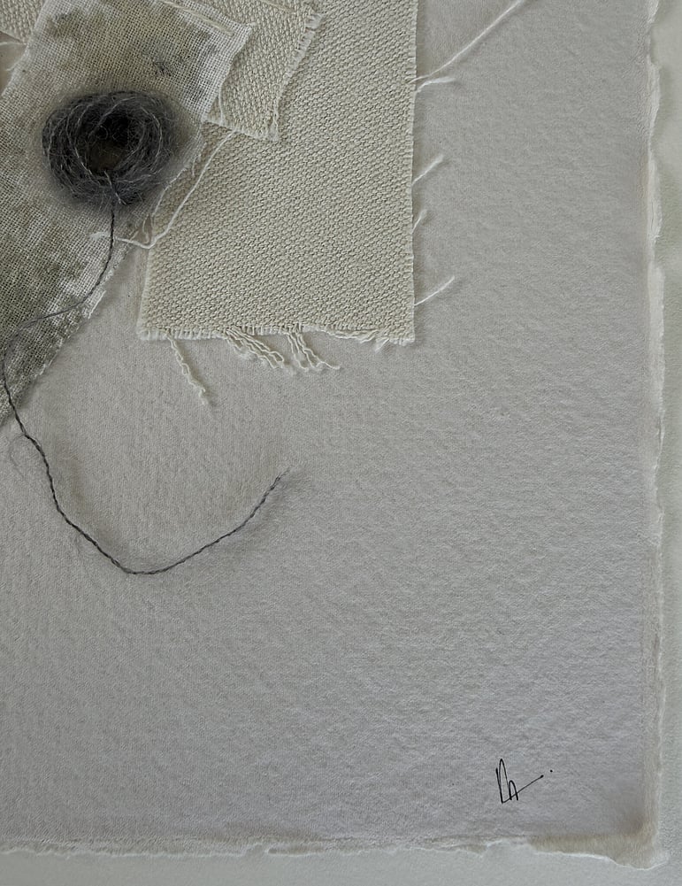 close-up image of sand-toned handmade cotton rag paper with deckled edge and visible surface texture, showing layered repurposed oat-toned linen and cotton fragments, one blotted with grey-green acrylic, soft frayed edges, charcoal baby alpaca/silk blend yarn remnant and handwritten signature in lower right corner 