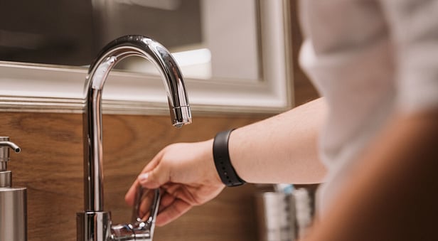 A person's hand reaching to adjust the lever on a modern, curved chrome faucet in a bathroom with a wooden backsplash.