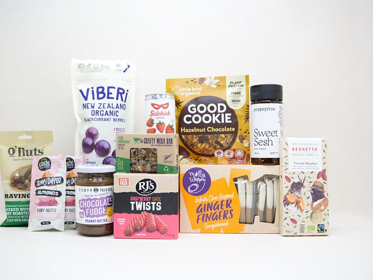 The Great NZ Snack Box featuring gourmet nuts, chocolate almonds, freeze-dried blackcurrants, strawberry soda, artisan chocolate, hot honey, cookies, muesli bars, raspberry twists, and Forty Thieves Chocolate Fudge Peanut Butter, packed with delicious New Zealand-made treats.