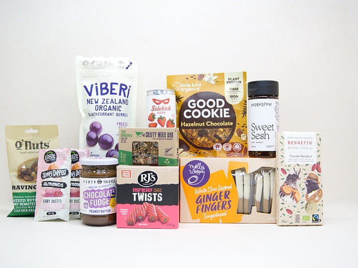 The Great NZ Snack Box featuring gourmet nuts, chocolate almonds, freeze-dried blackcurrants, strawberry soda, artisan chocolate, hot honey, cookies, muesli bars, raspberry twists, and Forty Thieves Chocolate Fudge Peanut Butter, packed with delicious New Zealand-made treats.