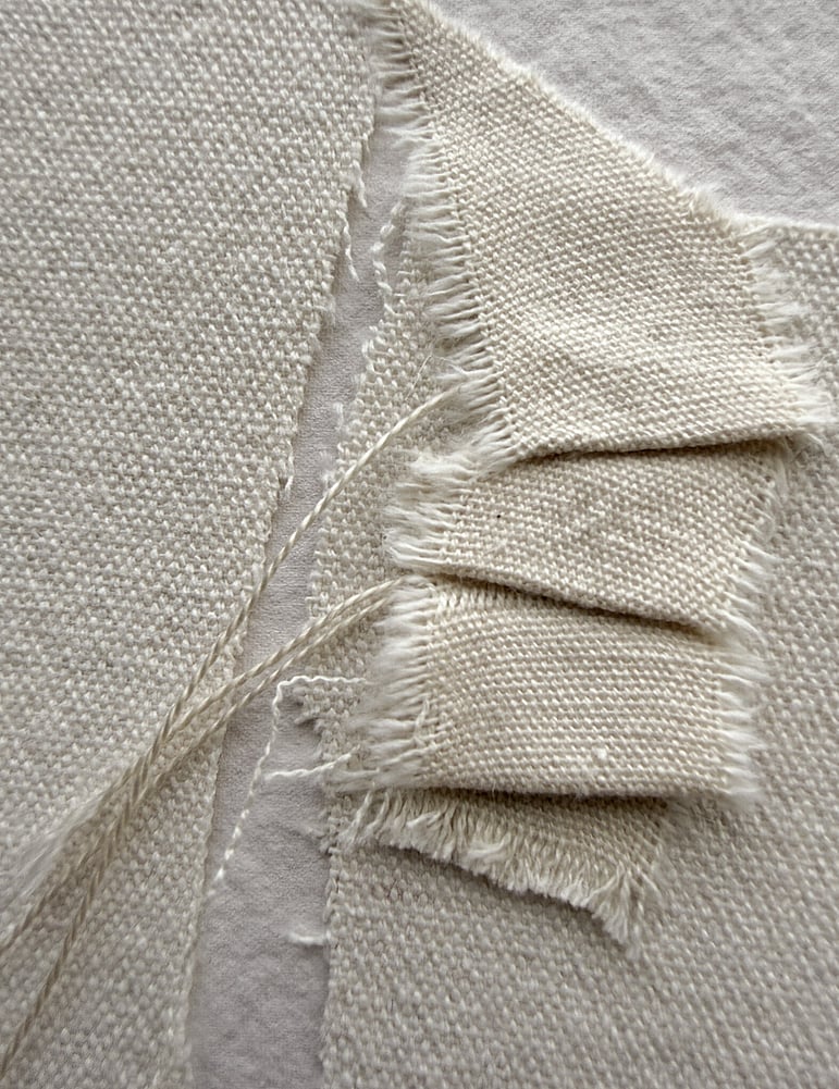 detail image of layered repurposed oat-toned cotton fragments and a small cotton fragment hand-stitched into gentle folds, showing visible weave texture, soft frayed edges and downward hanging threads 