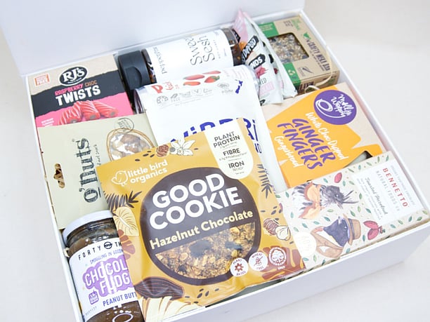 The Great NZ Snack Box featuring gourmet nuts, chocolate almonds, freeze-dried blackcurrants, strawberry soda, artisan chocolate, hot honey, cookies, muesli bars, raspberry twists, and Forty Thieves Chocolate Fudge Peanut Butter, packed with delicious New Zealand-made treats.