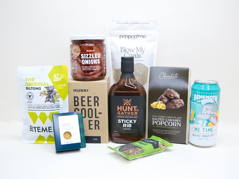 The BBQ Bloke Gift Box featuring Behemoth Me Time craft beer, a HUSKI beer cooler, Hunt & Gather Sticky Rib Sauce, Pepper & Me Blow My Coals seasoning, BiteMeat jerky, Chocolate Brown popcorn, Botanicals body bar, Pure Delish bar, and PICKLED sizzled onions, thoughtfully curated for BBQ lovers and craft beer fans.