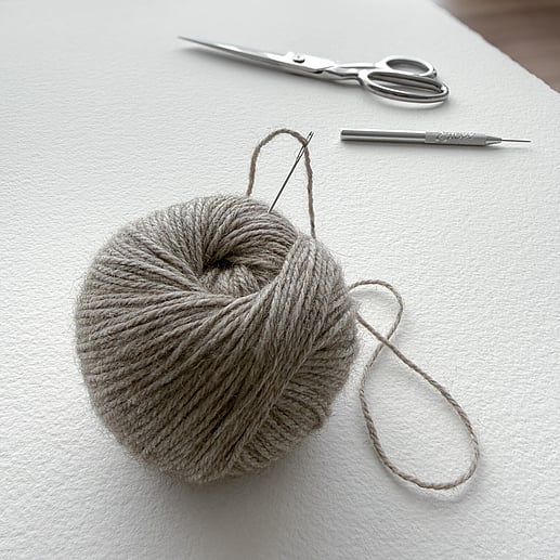 a skein of flax-toned wool threaded through a chenille needle resting on Arches fine art paper, with embroidery shears and a fine point awl in the background