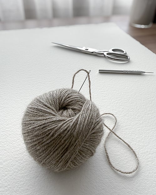image of a skein of flax-toned wool with a strand threaded through a chenille needle, resting on arches fine art paper with embroidery shears and a fine point awl in the background