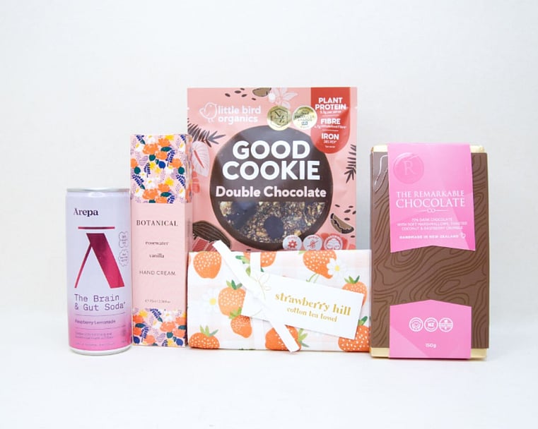The Pretty in Pink Gift Box featuring an Ārepa Raspberry Lemonade Brain Drink, Botanicals Rosewater & Vanilla Hand Cream, Little Bird Good Cookie, Strawberry Hill Tea Towel, and The Remarkables Marshmallow, Coconut & Raspberry Chocolate, beautifully curated for gifting in New Zealand.