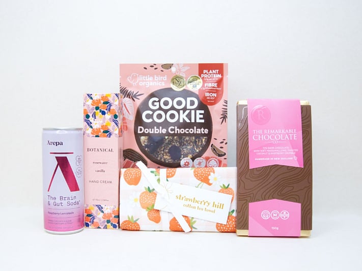 The Pretty in Pink Gift Box featuring an Ārepa Raspberry Lemonade Brain Drink, Botanicals Rosewater & Vanilla Hand Cream, Little Bird Good Cookie, Strawberry Hill Tea Towel, and The Remarkables Marshmallow, Coconut & Raspberry Chocolate, beautifully curated for gifting in New Zealand.