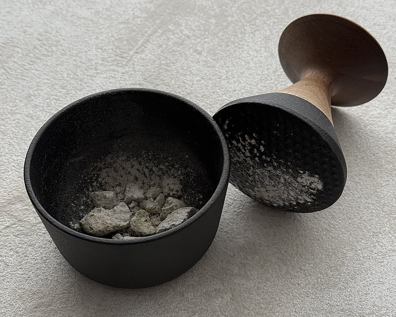process image showing hand-crushed pumice fragments and powder in a black mortar with pestle, used in 012 / 2026