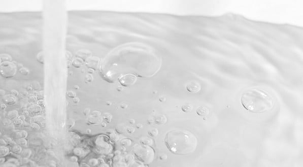Stream of clear water pouring into a basin, creating ripples and floating surface bubbles.