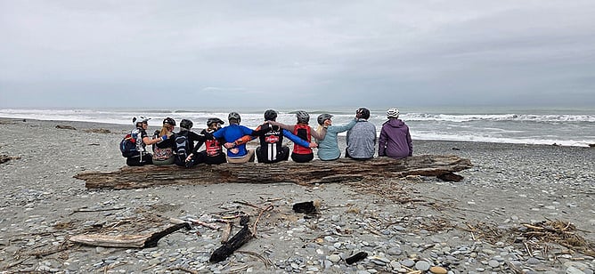 Cycle tour group resting on driftwood at a wild West Coast beach, West Coast Wilderness Trail, New Zealand