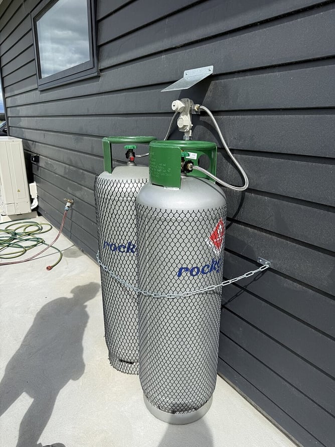 Two LPG gas bottles chained to a dark weatherboard wall and connected through a changeover regulator.