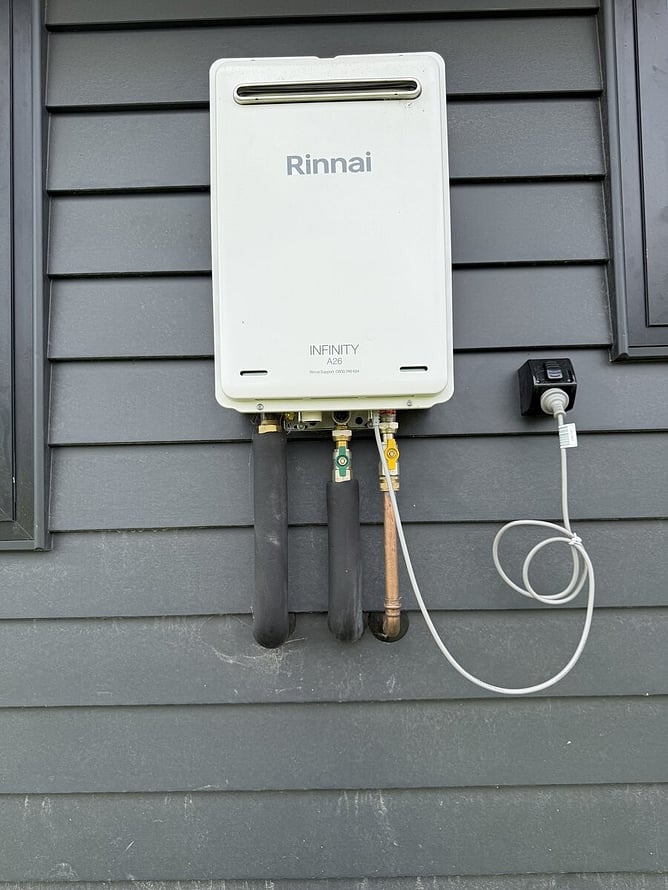 Rinnai Infinity continuous flow gas hot water unit mounted on an exterior grey weatherboard wall with its pipe connections and power plug.