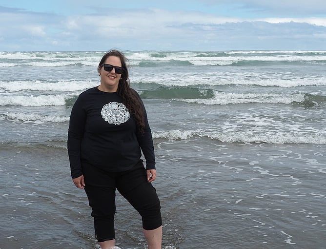 Anna Mollekin standing in the waves at Muriwai Beach wearing her seashell lace artwork tee