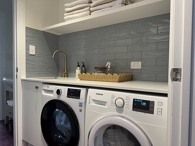 Modernized laundry room at Crab Shack Cottage in Russell featuring a front-loading washing machine, dryer, sink with brass tap, and a white subway tile splashback wall
