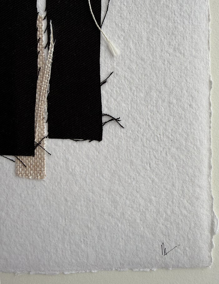 close-up image of handmade ivory-toned cotton rag paper with deckled edge and visible surface texture, two repurposed black cotton fragments with a slice of beige basketweave fabric wedged between them, varied weave texture, soft frayed edges, a trailing ecru thread remnant and handwritten signature in lower right corner 