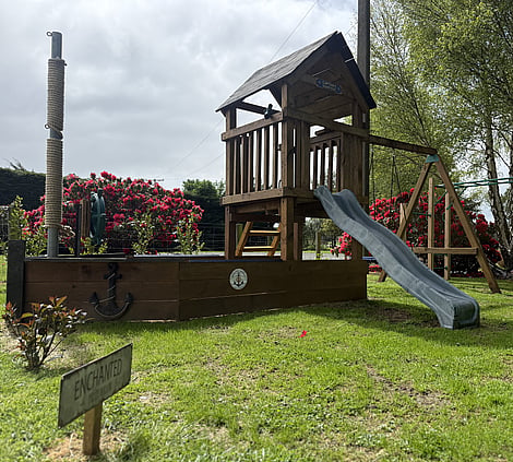 Play area at Forest Hillside Glamping Retreat, featuring a wooden children's playhouse with a slide, set in a spacious grassy garden surrounded by trees and colourful shrubs.