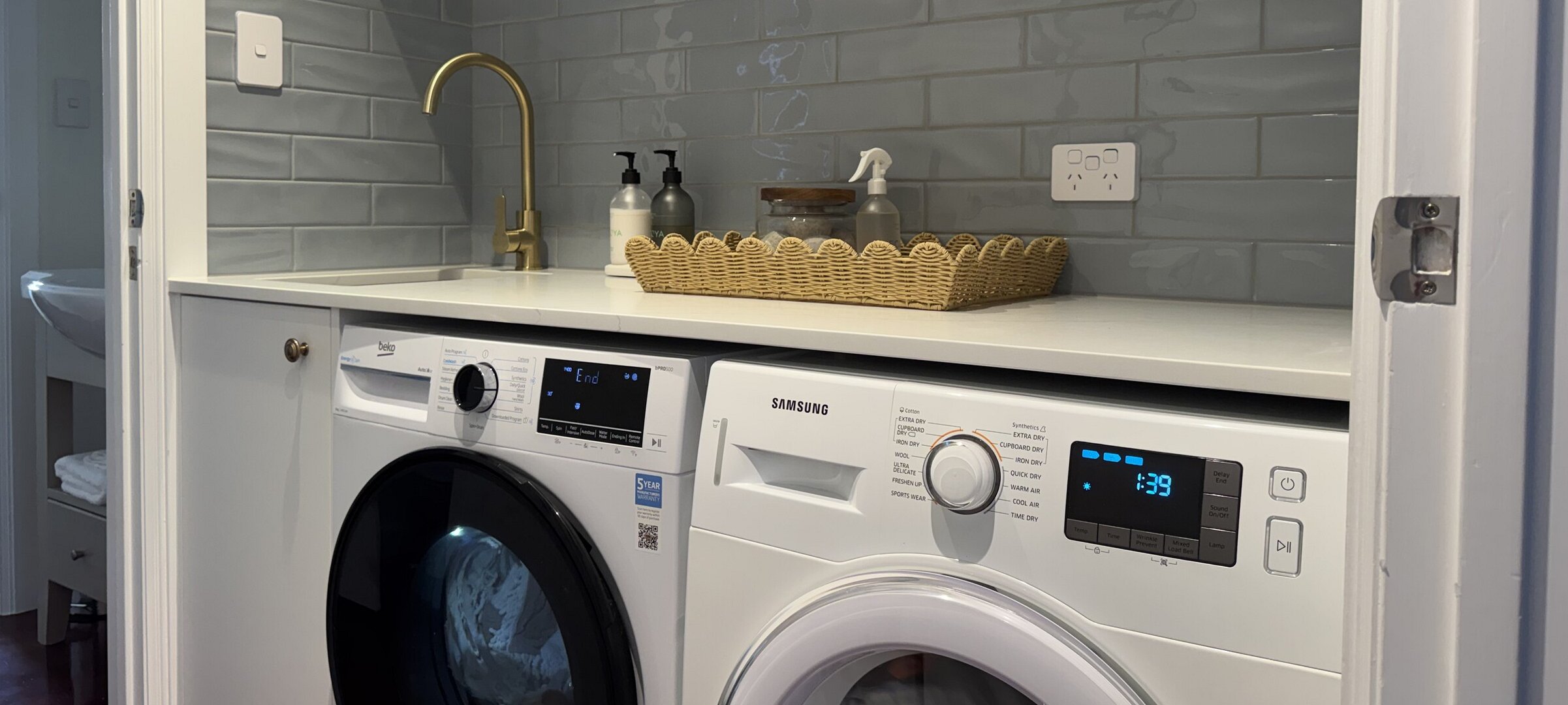 Modernised laundry room at Crab Shack Cottage in Russell featuring a front-loading washing machine, dryer, stainless steel sink, and white subway tile splashback wall