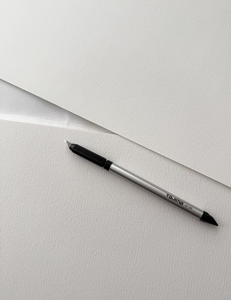 image of overlapping sheets of soft ivory fabriano fine art paper with a tajima art knife resting diagonally across the surface, in preparation for hand-cutting the pieces for c001 / 2026