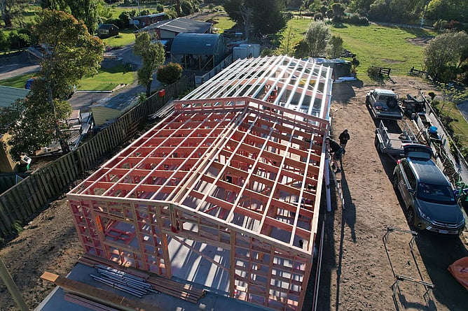 Aerial of the timber house framing and the steel garage frame standing on the slab in Kaiapoi, with the team and fleet alongside