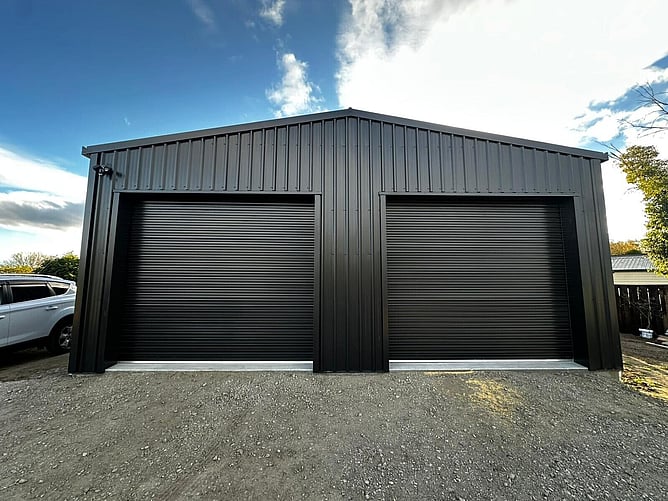 Front of the black garage and workshop with two roller doors, on a gravel driveway