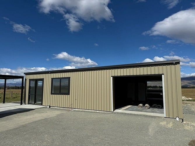 The finished cream shed home in Twizel with black joinery, the roller door open and a carport, under a big sky with mountains behind