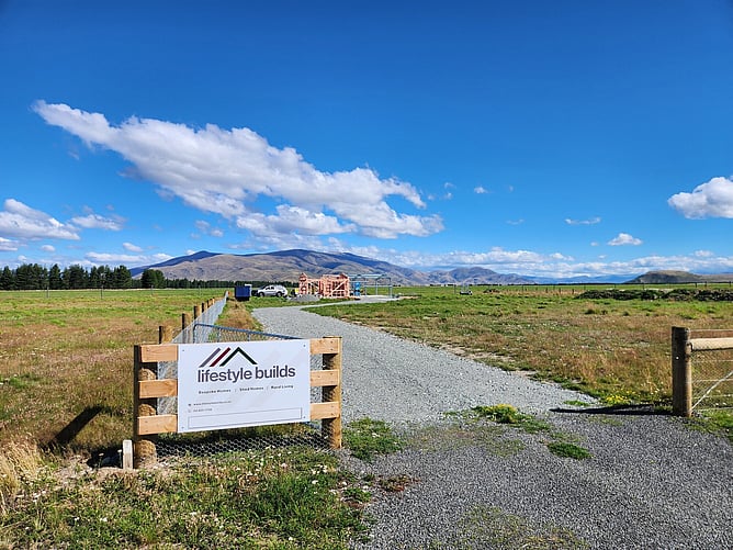 Lifestyle Builds sign at the gate with the gravel drive leading to the frame and the mountains beyond
