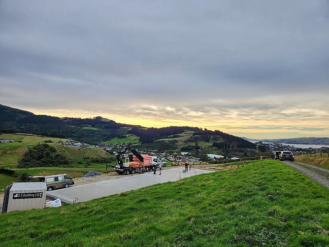 The poured slab on the Dunedin hillside with a Hiab truck unloading the kitset, DS Building's site container in the foreground and the town and water below