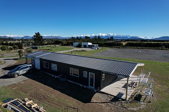 Aerial of the black Oxford shed home with the snow-covered Southern Alps behind and the horse arena next door