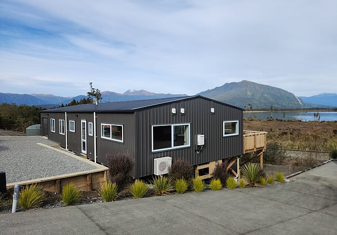 The finished four-bedroom bach on its timber piles above Lake Brunner, with the mountains behind