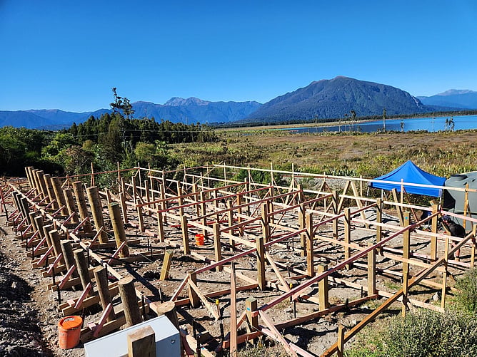 Rows of timber piles and bearers being set out for the Moana bach, with Lake Brunner and the mountains behind