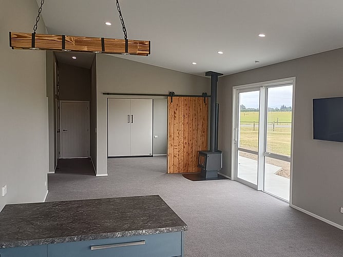 The living room with a wood burner, timber barn door and a hanging timber beam light, paddocks outside the glass