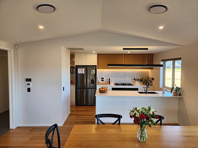 Kitchen and dining under the raking ceiling, with a herringbone splashback, timber overhead cupboards and a black linear pendant