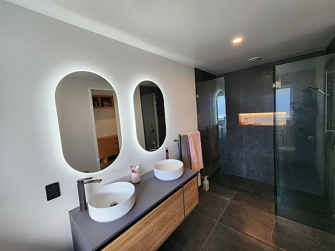 The tiled ensuite in the Rolleston showhome: double vessel basins, backlit oval mirrors and the twin-head shower behind glass