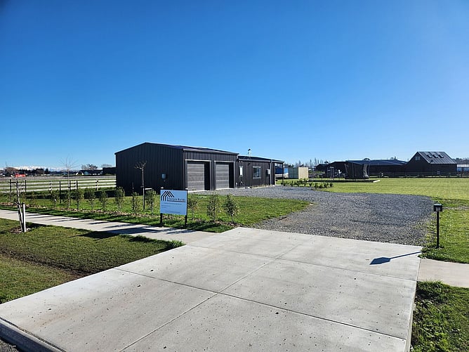 The finished Lincoln shed home from the road: black steel cladding, two roller doors, a gravel drive and the Lifestyle Builds sign out front